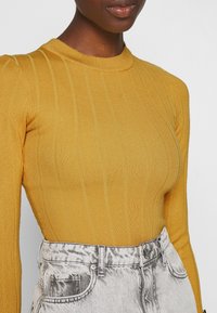 Hořčicově žlutý žebrovaný top s dlouhým rukávem a kulatým výstřihem, v kombinaci s vysokým pasem stříbrných džínů ze denimu s opotřebovanými detaily.