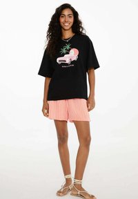 Žena s dlouhými kudrnatými vlasy, oblečená v černém tričku s potiskem, oranžových a bílých pruhovaných šortkách a bílých sandálech na tkaničky, se usmívá a stojí.