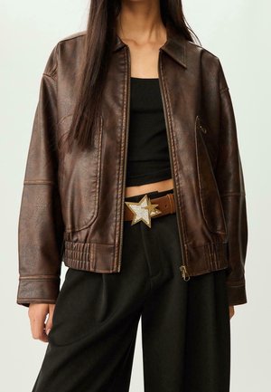 Kobieta ubrana w brązową skórzaną kurtkę, czarny krótki top, czarne spodnie i brązowy pasek z dużą, gwiazdzistą klamrą.