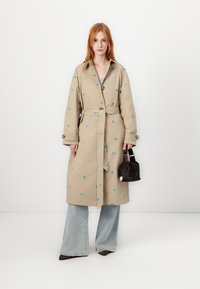 Beige trenchcoat met groene bloemenborduring, knoopsluiting aan de voorkant, ceintuur in de taille, zijzakken, gecombineerd met flare jeans en een donkere handtas.
