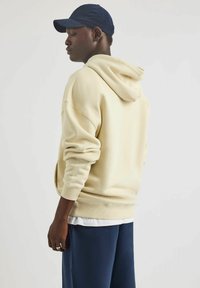 Kremowa bluza z luźnym krojem, z kapturem ze sznurkiem i ściągaczami na rękawach, zestawiona z granatowymi spodniami. Gładka faktura, casualowy design.