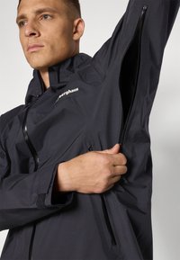 Black lightweight jacket with a zippered chest pocket, high collar, and textured fabric. Features a small white logo on the left side.