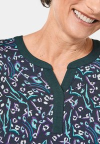 Kurzärmliges Oberteil mit V-Ausschnitt, aus einem dunklen Stoff mit einem bunten, abstrakten Druck in Lila, Türkis und Weiß.