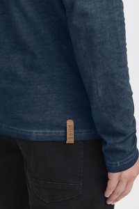 Langarm-Hemd in Marineblau aus strukturiertem Stoff, ausgestattet mit einem Lederetikett mit dem Wort "solid" und einem geraden Saum. Kombiniert mit dunklen Hosen.