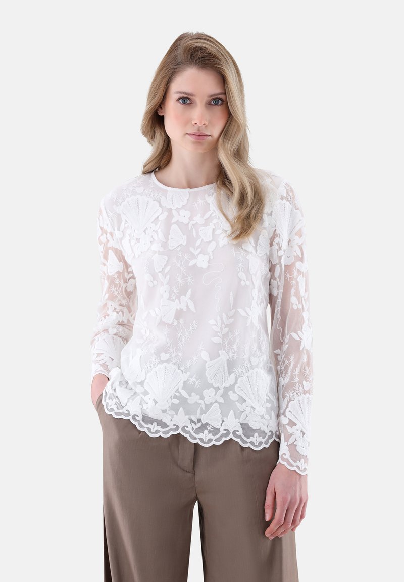 Weiße, durchsichtige Bluse mit floraler Stickerei und gewelltem Saum. Lange Ärmel und runder Ausschnitt. Über beigen Hosen getragen.