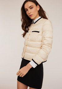 Giacca puffer color crema, con zip frontale, dettagli neri e due tasche nere. Indossata sopra una camicia bianca con colletto e una gonna nera.