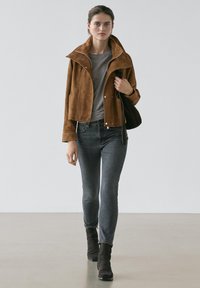 Kvinna som går och bär en brun mockajacka, grå t-shirt, urtvättade svarta jeans, svarta stövlar och bär en svart axelväska mot en enfärgad bakgrund.