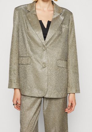 Kvinde iført en metallisk guldtekstureret dragt med en dobbeltknappet blazer og matchende bukser over en sort top.