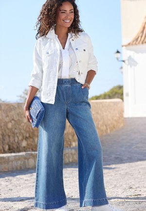 Woman standing outdoors, wearing white jacket, embroidered blouse, wide-leg blue jeans, holding blue woven clutch, smiling against stone wall background.
