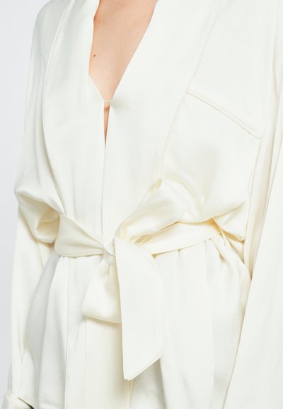 Κρέμα μπλούζα με τύλιγμα και βαθύ V-ντεκολτέ, μακριά μανίκια και ζώνη που δένει μόνη της. Φτιαγμένη από μαλακό, ελαφρύ ύφασμα.