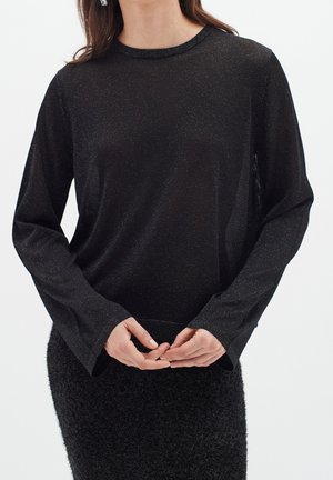Woman wearing a black long-sleeve glittery top and textured black skirt, standing with hands clasped in front against a white background.