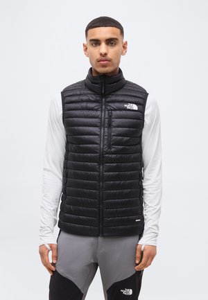 Uomo che indossa un gilet trapuntato nero di North Face sopra una camicia bianca a maniche lunghe e pantaloni grigi neri, in piedi davanti a uno sfondo semplice.