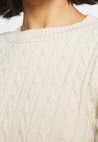 Beiger, kabelgestrickter Pullover mit einem gerippten Rundhalsausschnitt. Die Textur ist weich mit ausgeprägten verdrehten Mustern und dezentem Glanz.