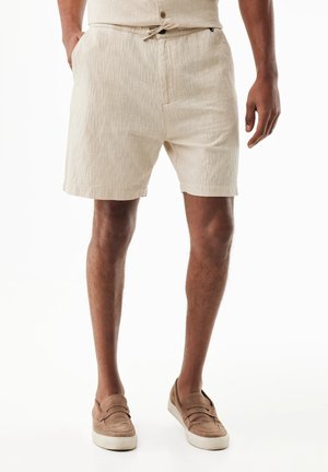 Mann trägt beige strukturierte Shorts und hellbraune Slip-On-Schuhe, steht mit teilweise in den Taschen steckenden Händen vor weißem Hintergrund.