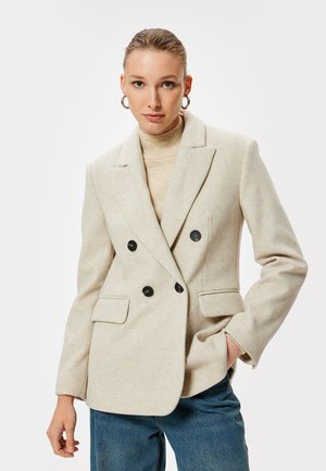 Blazer beige doppiopetto realizzato in misto lana, con bottoni neri, due tasche anteriori e rever a pico. Indossato sopra un dolcevita color cammello.