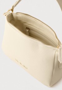 Crèmewitte crossbody tas van getextureerd leer met ritssluiting, voorzien van een goudkleurig logo en een ritsvak aan de binnenkant.