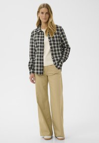 Donna con lunghi capelli biondi, che indossa una camicia a quadri bianca e nera, un top bianco, pantaloni larghi beige e scarpe bianche, in piedi contro uno sfondo bianco.