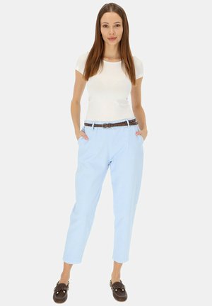 Jasnoniebieskie spodnie damskie z brązowym paskiem, biały top z krótkim rękawem oraz brązowe mokasyny, stojąca z rękami w kieszeniach na białym tle.