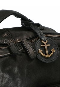 Borsa in pelle nera con un charm ad ancora in bronzo su un'etichetta circolare, con cuciture visibili e dettagli della cerniera. Texture liscia in tutto.