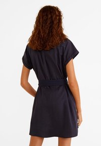 Robe courte bleu marine avec une ceinture, des manches courtes et une coupe décontractée. Présente un design simple avec une texture de tissu lisse.