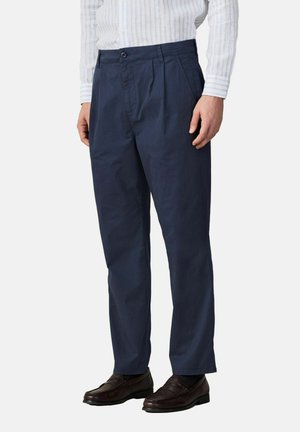 Mann trägt marineblaue plissierte Hose, weiß gestreiftes langärmliges Hemd und dunkelbraune Lederloafer, steht vor einem schlichten Hintergrund.