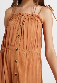 Kvinna som bär en ärmlös, roströd knapar-kjolklänning med volangöverkant och tunna axelband.