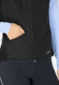 Schwarze gepolsterte Weste mit einem Frontreißverschluss, strukturierten Oberflächen und einer Tasche. Kombiniert mit einem hellblauen Langarmoberteil und dunklen Leggings.