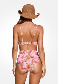 Różowy bikini w kwiaty z cienkimi ramiączkami, z wysokim stanem i szczegółowym wzorem zielonego liścia. Noszone z słomkowym kapeluszem przeciwsłonecznym.