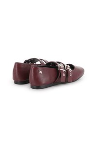 Un par de zapatos planos burdeos con puntera afilada, correas dobles con hebillas plateadas, suelas negras y un pequeño logotipo metálico en el talón.