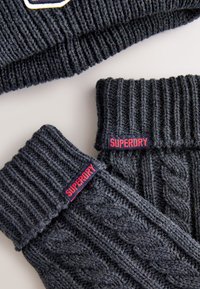 Donkergrijze gebreide handschoenen en een bijpassende muts met geribbelde en kabelpatronen, met opvallende rode "SUPERDRY"-labels op elk stuk.