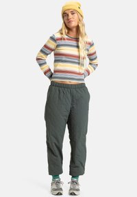 Gestreepte lange mouw top in geel, blauw en rood; groene elastische broek; grijze sportschoenen; geribbeld gele beanie.