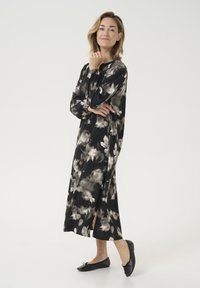 Vestido largo negro con estampado floral blanco, tela suave, escote redondo, puños elásticos y aberturas laterales. Lleva zapatos planos negros.