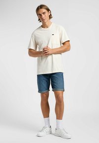 Camiseta corta de manga corta en color crema con un bolsillo en el pecho, combinada con pantalones cortos de mezclilla enrollados y zapatillas blancas, mostrando calcetines blancos visibles por encima de los zapatos.