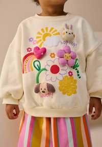 Sweatshirt couleur crème avec des motifs brodés colorés, y compris des fleurs, un soleil, un arc-en-ciel et des motifs d'animaux. Coupe décontractée et texture douce.
