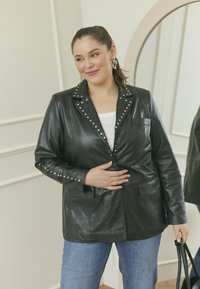 Schwarze Lederblazer mit Nietenkragen und -manschetten, ausgestattet mit einer einzigen Brusttasche und zwei vorderen Taschen, getragen über einem weißen Oberteil.
