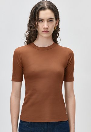 Mladá žena s kučeravými tmavými vlasmi, ktorá má na sebe zlaté kruhové náušnice, priliehavý hnedý krátky rukáv a tmavomodré džínsy, stojaca pred sivým pozadím.