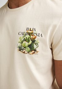 Cremefarbenes Baumwoll-T-Shirt mit einem bunten Kakteen-Motiv und dem Text "BAJA CALIFORNIA MX" in Schwarz, zentral auf der Brust positioniert.