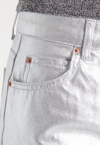 Stříbrné metalické džíny s hladkou texturou, designem s pěti kapsami, měděnými nýty a vysokým pasem. Viditelné švy.