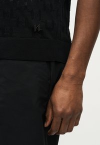 Χέρι σκουρόχρωμου ατόμου ακουμπισμένο στο πλάι, φορώντας μαύρο πλεκτό τοπ με υφή και μικρό μαύρο διακριτικό λογότυπο καθώς και μαύρο παντελόνι.