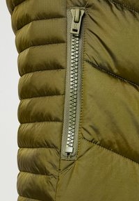 Zielona oliwkowa kamizelka puchowa z lekko błyszczącego, pikowanego materiału, pionowymi przeszyciami i kontrastowym zamkiem błyskawicznym z uchwytem.