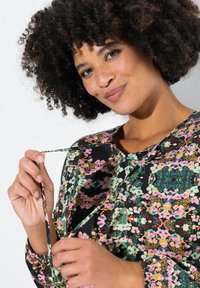 Flerfärgad blus med mönster på svart botten, med blommotiv och långa ärmar. Modellen håller en matchande knytning nära halsringningen.