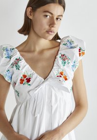 Vit klänning med puffärmar, prydd med blommig broderi i blått, rött och orange. Knytning vid halslinningen och plisserad byst.