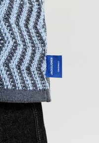 Tissu tricoté en zigzag bleu clair et gris avec une étiquette bleue portant la mention "Jack & Jones Originals" au-dessus d'un jean en denim foncé.