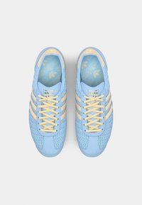 Zapatillas de color azul claro con un upper texturizado, tres franjas amarillas y cordones a juego. Presenta un logo dorado en la lengüeta y una plantilla con patrón.