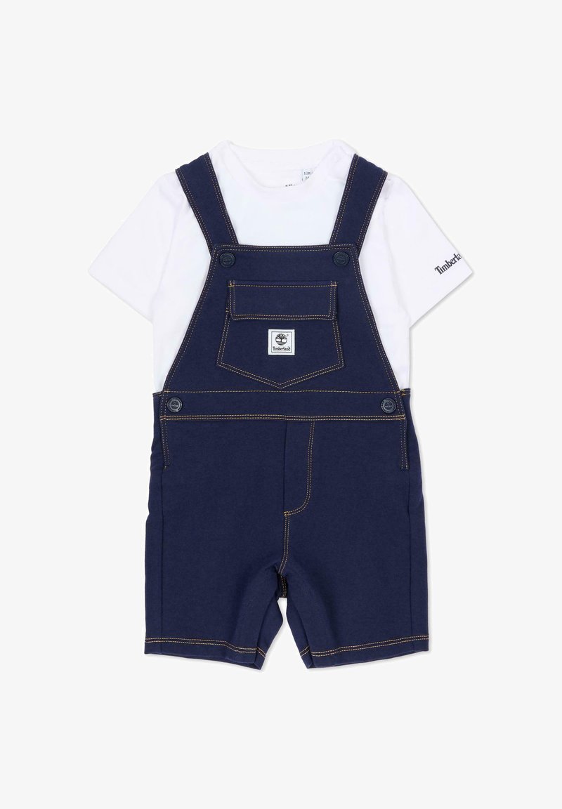 Salopette en denim bleu marine à jambes courtes, avec une poche frontale, des bretelles réglables et des coutures dorées. Associée à un t-shirt blanc en dessous.