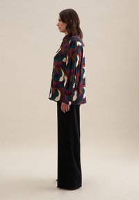 Blusa colorata a fantasia con forme curve in viola, verde e crema; maniche lunghe e chiusura a bottone, abbinata a pantaloni neri a gamba larga.