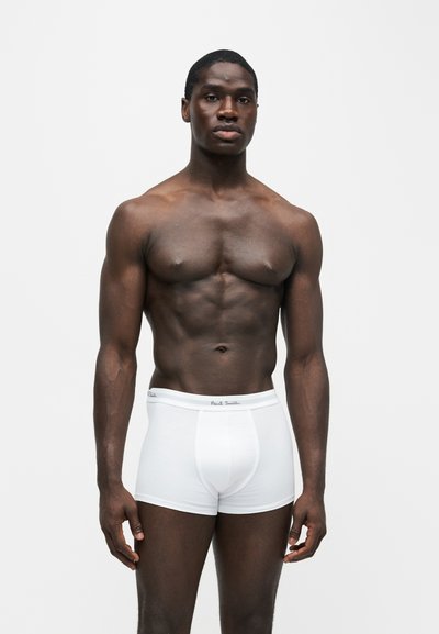 Homme musclé à la peau foncée debout face à nous, portant un boxer blanc Paul Smith sur un fond clair uni.