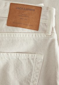 Bež traper tkanina s kožnom etiketom na kojoj piše "JACK & JONES", smještenom iznad stražnjeg džepa. Tekstura je glatka s vidljivim šavovima.