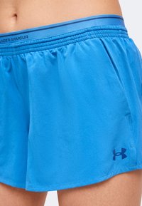 Μπλε αθλητικό σορτς Under Armour φορεμένο στη λεκάνη και το πάνω μέρος του μηρού ενός ατόμου με ελαστική μέση και λογότυπο στο μπροστινό κάτω μέρος.