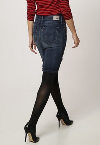 Jeanskjol i mörk tvätt, figurnära modell, medellång, med signatur bakfickor. Bärs med svarta högklackade skor och genomskinliga svarta strumpbyxor.